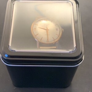 May include: A vintage gold-toned wristwatch with a light-colored face, hands, and hour markers, displayed in a black box with a clear lid. The watch face has the word "HAMILTON" printed on it.