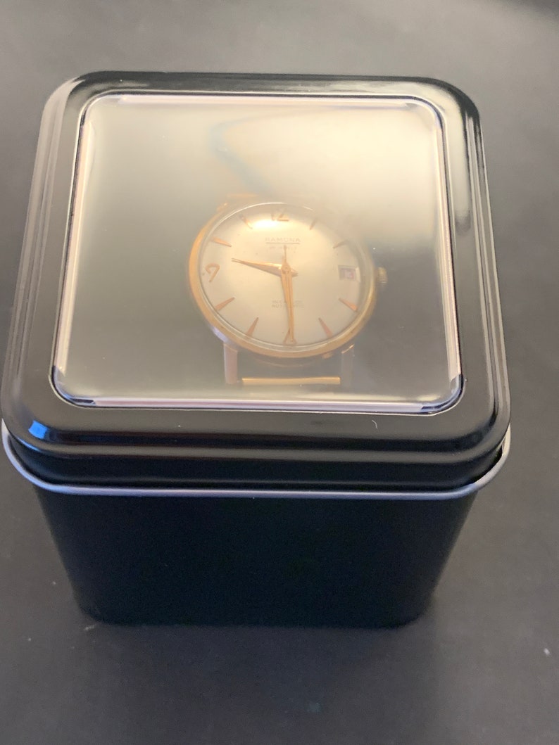 May include: A vintage gold-toned wristwatch with a light-colored face, hands, and hour markers, displayed in a black box with a clear lid. The watch face has the word "HAMILTON" printed on it.
