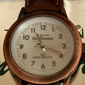 K&ouml;nnte beinhalten: Nahaufnahme einer Ben Sherman-Uhr mit braunem Lederarmband. Das Zifferblatt ist cremefarben mit schwarzen Ziffern und Zeigern. Das Uhrengeh&auml;use hat eine Kupferoberfl&auml;che. Der Text "50M Water Resistant" ist sichtbar.