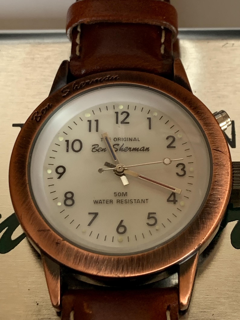 K&ouml;nnte beinhalten: Nahaufnahme einer Ben Sherman-Uhr mit braunem Lederarmband. Das Zifferblatt ist cremefarben mit schwarzen Ziffern und Zeigern. Das Uhrengeh&auml;use hat eine Kupferoberfl&auml;che. Der Text "50M Water Resistant" ist sichtbar.