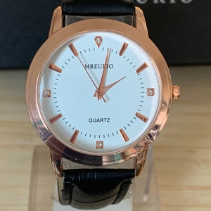 Puede incluir: Un reloj de oro rosa con una esfera blanca y una correa de cuero negro. La esfera del reloj tiene un diamante en la posición de las 12 en punto y las palabras "MREUTIO" y "QUARTZ" impresas en ella.