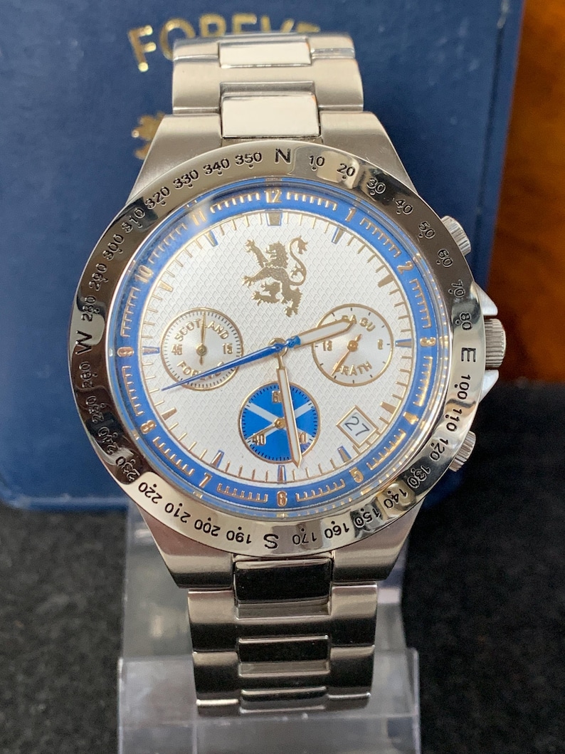 Puede incluir: Reloj de pulsera plateado con esfera blanca y detalles azules. El reloj presenta m&uacute;ltiples subesferas, una pantalla de fecha y un bisel giratorio con marcas de br&uacute;jula. La correa es de metal plateado.