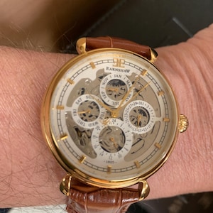May include: A gold-tone wristwatch with a brown leather strap featuring a textured pattern. The watch face is cream-colored with a skeleton design, multiple sub-dials for the day, month, and date, and the text EARNSHAW 1805 printed on the dial.