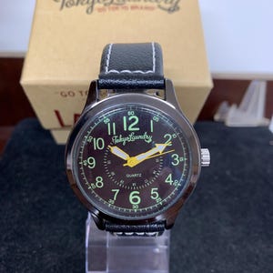 May include: A black wristwatch with a black leather band and a green face. The face has white numbers and hands, and the words "Tokyo Laundry" in yellow. The watch says "Quartz" below the 12.
