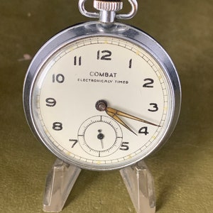 Puede incluir: Un reloj de bolsillo plateado con una esfera blanca y agujas negras. El reloj tiene una caja cromada y una corona negra. La esfera del reloj dice "COMBAT ELECTRONICALLY TIMED" y tiene números negros del 1 al 12. El reloj está en un soporte plateado.