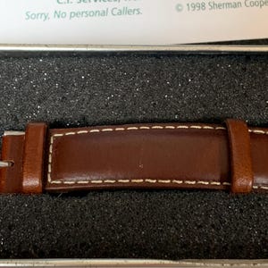 K&ouml;nnte beinhalten: Nahaufnahme eines braunen Leder-Uhrenarmbands mit wei&szlig;en N&auml;hten. Das Armband befindet sich in einer dunklen, strukturierten Box. Das Uhrenarmband hat eine silberne Schnalle und ist wahrscheinlich Teil einer Vintage- oder klassischen Uhr.