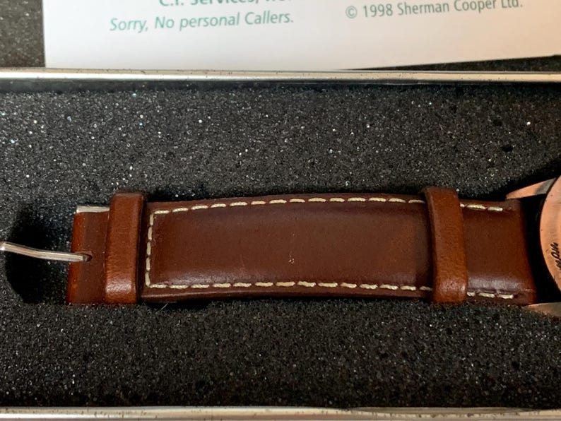 K&ouml;nnte beinhalten: Nahaufnahme eines braunen Leder-Uhrenarmbands mit wei&szlig;en N&auml;hten. Das Armband befindet sich in einer dunklen, strukturierten Box. Das Uhrenarmband hat eine silberne Schnalle und ist wahrscheinlich Teil einer Vintage- oder klassischen Uhr.