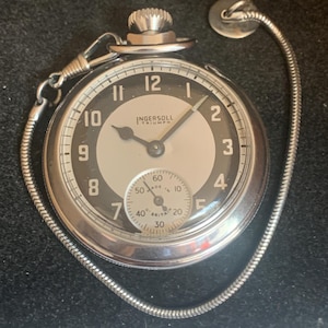 May include: A silver-toned Ingersoll Triumph pocket watch with a round face featuring a black outer ring, a white center, and black Arabic numerals. It includes a small sub-dial for seconds, a winding crown at the top, and a metal chain attached.