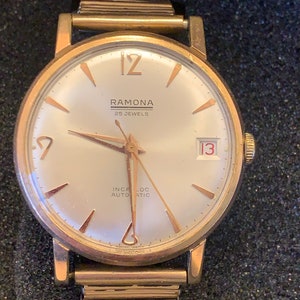 May include: A gold-toned Ramona wristwatch with a light-colored face. The watch features gold hands and markers, a date window at the 3 o'clock position, and the text "RAMONA 25 JEWELS INCA-LOC AUTOMATIC". The watch band is also gold-toned.