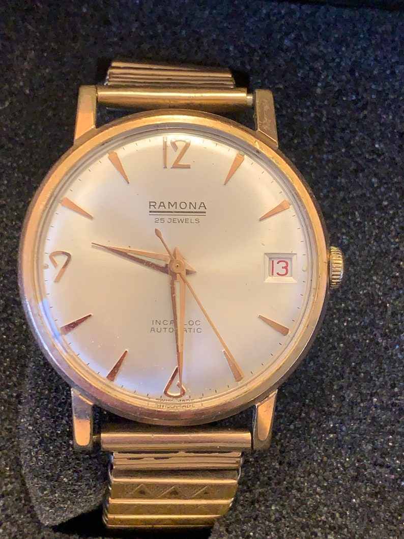 May include: A gold-toned Ramona wristwatch with a light-colored face. The watch features gold hands and markers, a date window at the 3 o'clock position, and the text "RAMONA 25 JEWELS INCA-LOC AUTOMATIC". The watch band is also gold-toned.
