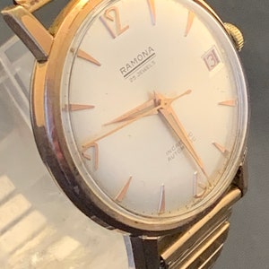 May include: A vintage gold-toned Ramona wristwatch with a white face, gold hands, and hour markers. The watch face displays the text "RAMONA 25 JEWELS" and a date window. The watch has a gold-toned band.