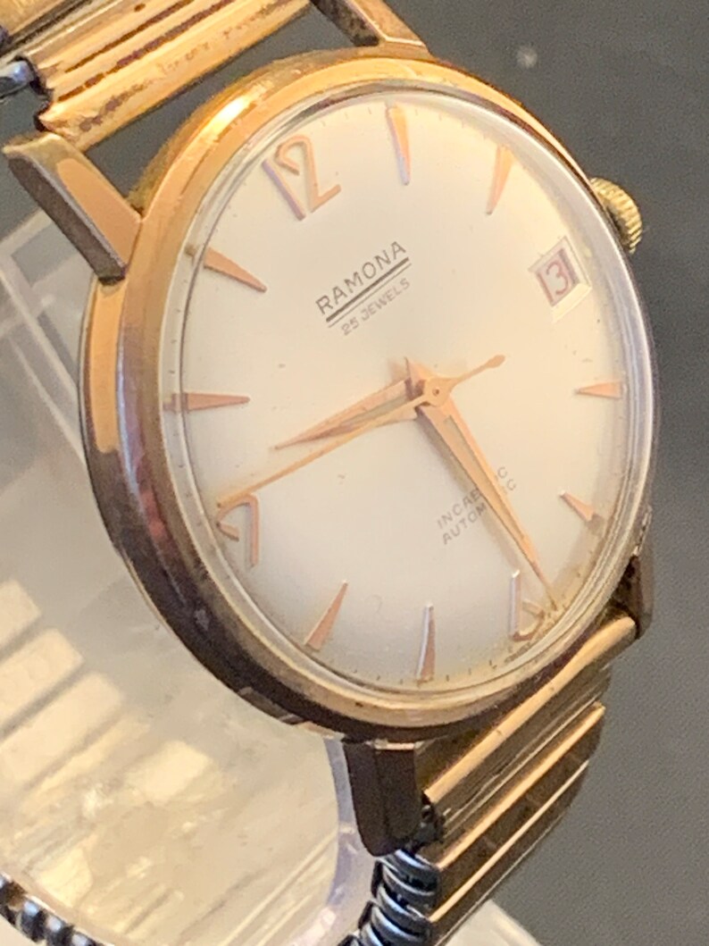 May include: A vintage gold-toned Ramona wristwatch with a white face, gold hands, and hour markers. The watch face displays the text "RAMONA 25 JEWELS" and a date window. The watch has a gold-toned band.