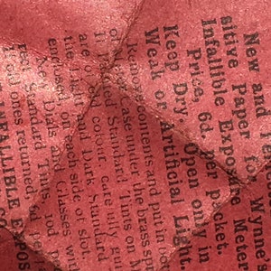 Op de afbeelding: Een close-up van een gekreukeld, rood papieren fragment met zwarte tekst. De tekst bevat zinnen als "New and Perfected Sensitive Paper" en "Infallible Exposure Meter". Het papier lijkt verouderd en versleten.