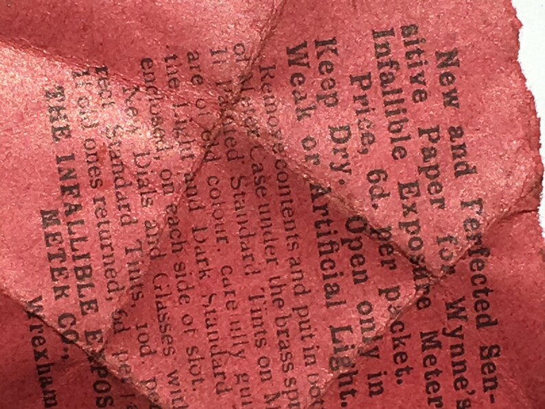 Op de afbeelding: Een close-up van een gekreukeld, rood papieren fragment met zwarte tekst. De tekst bevat zinnen als "New and Perfected Sensitive Paper" en "Infallible Exposure Meter". Het papier lijkt verouderd en versleten.