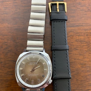 Puede incluir: Un reloj vintage Kay Time Super De Luxe con una correa de metal plateado y una correa de cuero negro separada. La esfera del reloj es marrón con marcadores y agujas plateadas. El texto "WATER RESISTANT ANTIMAGNETIC" es visible.