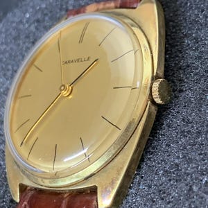 May include: A gold-toned wristwatch with a brown leather band. The watch face is round and has a gold background with black markings. The word "CARAVELLE" is printed in black on the watch face.