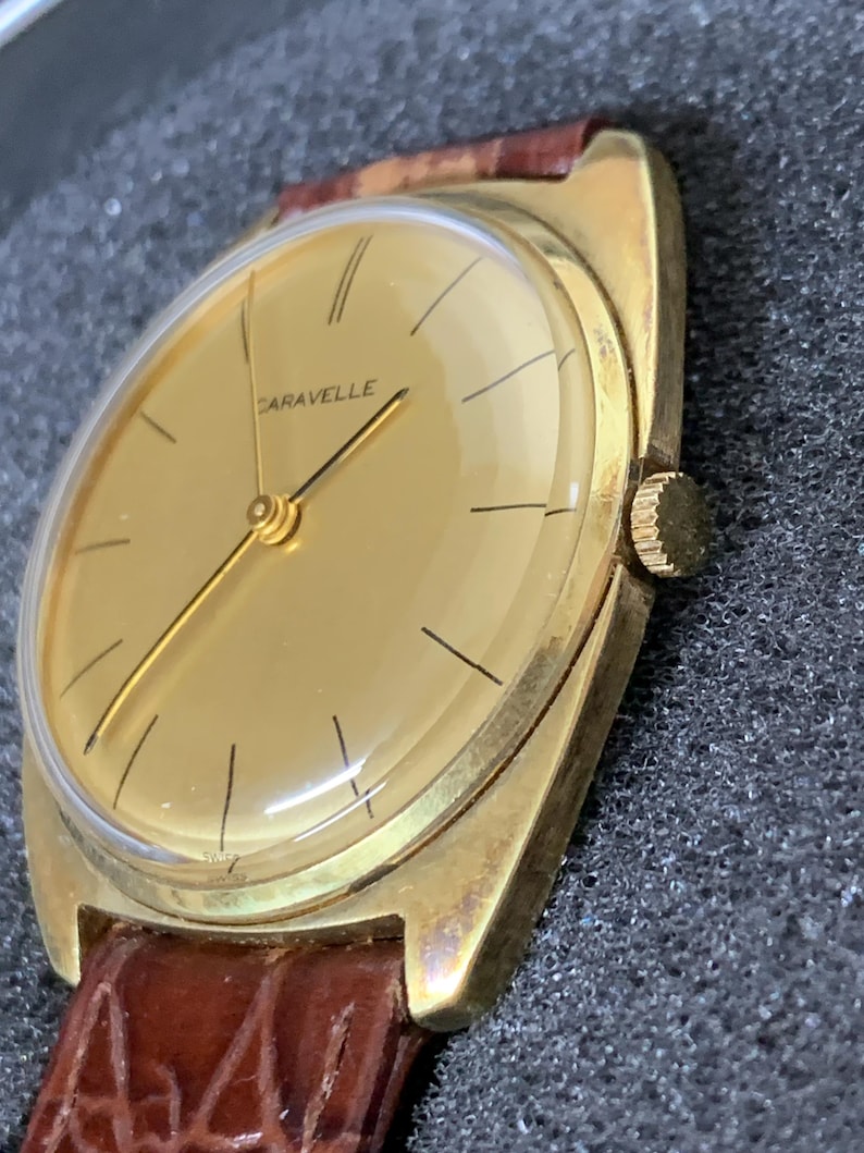 May include: A gold-toned wristwatch with a brown leather band. The watch face is round and has a gold background with black markings. The word "CARAVELLE" is printed in black on the watch face.