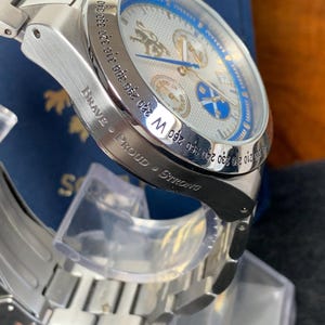 Puede incluir: Reloj de pulsera plateado con esfera azul y blanca, con m&uacute;ltiples subesferas y un bisel estilo br&uacute;jula. La correa del reloj tambi&eacute;n es plateada. Las palabras "Brave - Proud - Strong" est&aacute;n grabadas en el lateral. El reloj tiene un dise&ntilde;o detallado.