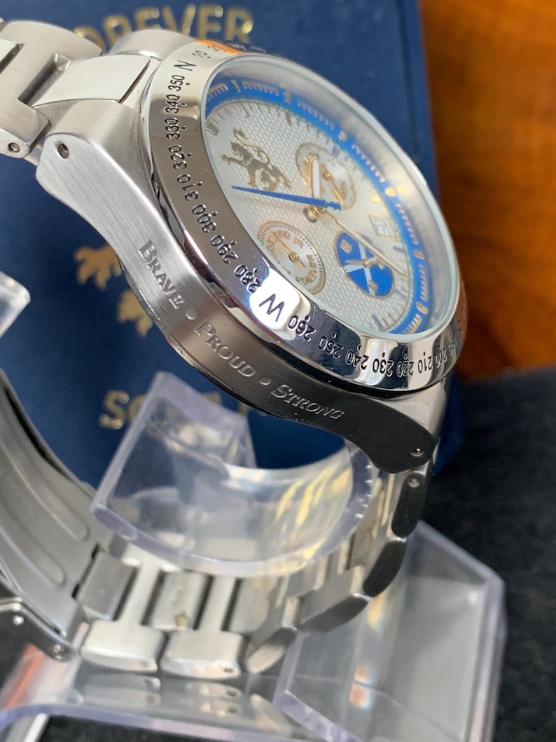 Puede incluir: Reloj de pulsera plateado con esfera azul y blanca, con m&uacute;ltiples subesferas y un bisel estilo br&uacute;jula. La correa del reloj tambi&eacute;n es plateada. Las palabras "Brave - Proud - Strong" est&aacute;n grabadas en el lateral. El reloj tiene un dise&ntilde;o detallado.