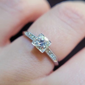 May include: A silver engagement ring with a square-shaped diamond set in a prong setting. The band is adorned with small diamonds.