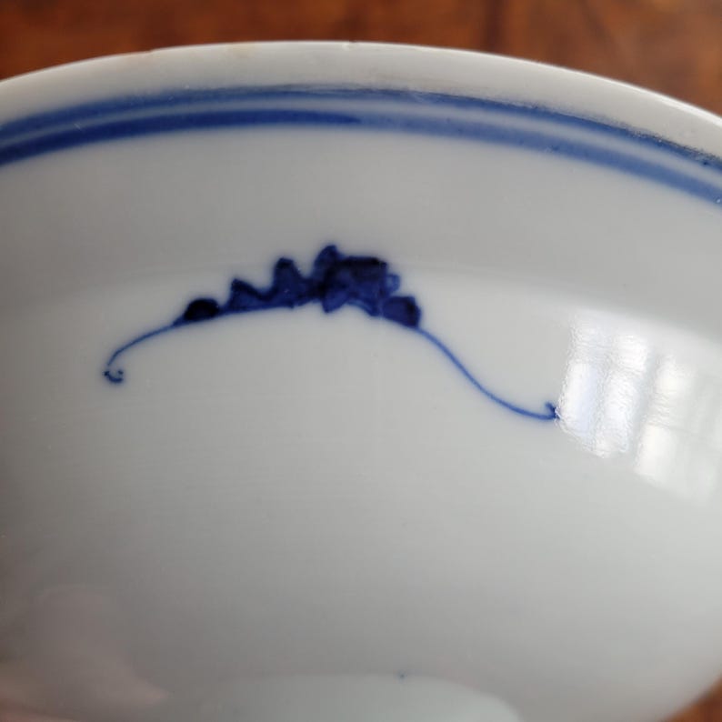 May include: White ceramic bowl with a blue floral design and a blue stripe around the rim. The bowl has a smooth, glossy finish and a simple, elegant design. The blue floral design is hand-painted.