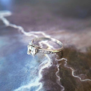 May include: A silver engagement ring with a single round diamond set in a square prong setting. The band is decorated with small diamonds.