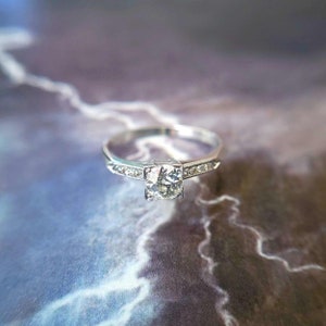 May include: A silver engagement ring with a single, round, clear diamond in the center. The band is set with smaller diamonds.