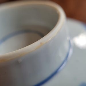 May include: Close-up of a white ceramic teacup with a blue painted design. The rim of the cup has a slight yellowed discoloration. The cup is sitting on a matching saucer, with a blue painted design.
