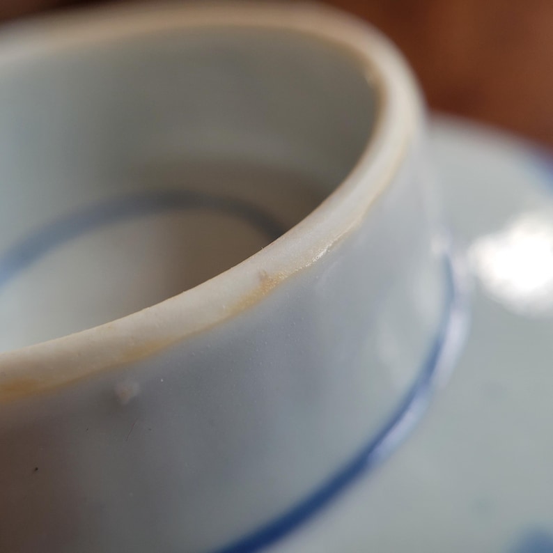 May include: Close-up of a white ceramic teacup with a blue painted design. The rim of the cup has a slight yellowed discoloration. The cup is sitting on a matching saucer, with a blue painted design.