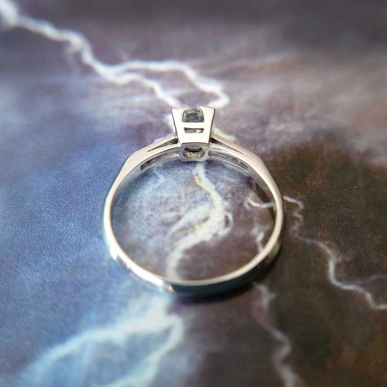 May include: A silver engagement ring with a single round diamond set in a prong setting. The ring is shown from the side, with the diamond facing the camera.