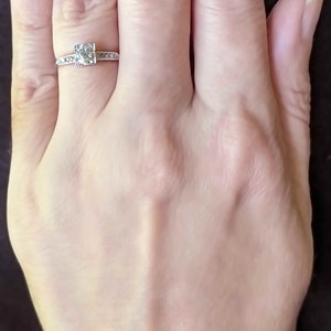 May include: A close-up of a hand wearing a silver ring with a single, round, clear gemstone surrounded by smaller gemstones. The ring is on the ring finger of the left hand.