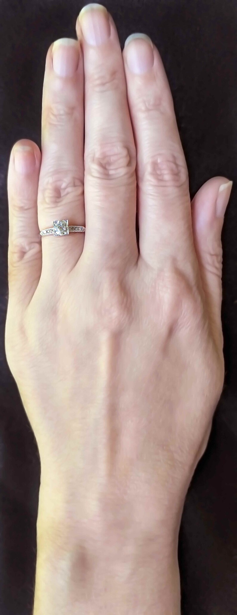May include: A close-up of a hand wearing a silver ring with a single, round, clear gemstone surrounded by smaller gemstones. The ring is on the ring finger of the left hand.