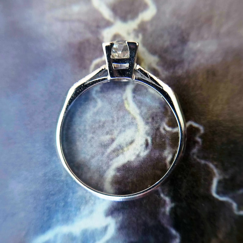 May include: A silver ring with a clear gemstone set in a prong setting. The ring is on a marble-like surface.