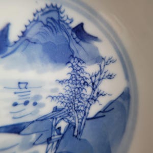 May include: A close-up of a white ceramic plate with a blue landscape design. The design features mountains, trees, and a small structure near water. The plate's rim has a blue border.