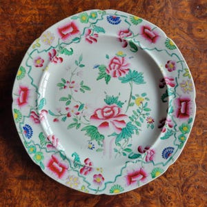 May include: A round ceramic plate with a scalloped edge featuring a white base decorated with hand-painted pink and red floral motifs, green leaves, and small blue and yellow flower accents arranged in a central cluster and along the rim.