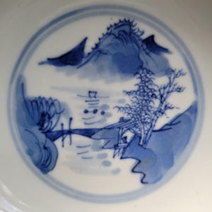 May include: A white ceramic plate with a blue landscape design. The design features a mountain, trees, and a body of water. The plate has a blue border and the landscape is painted in shades of blue.