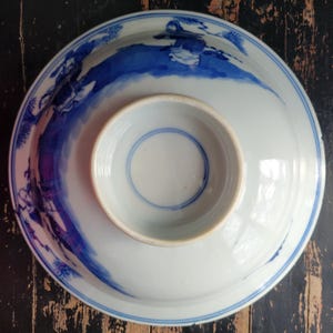 May include: A white porcelain bowl with a blue painted landscape design. The bowl has a circular base with a smaller, raised circular center. The rim of the bowl and the center have blue painted lines.