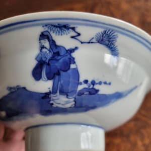 May include: A white porcelain bowl with a blue painted design. The bowl features a detailed illustration of a figure in traditional clothing, set against a landscape with trees. The rim is accented with a blue border. The bowl is elevated on a short, white pedestal.