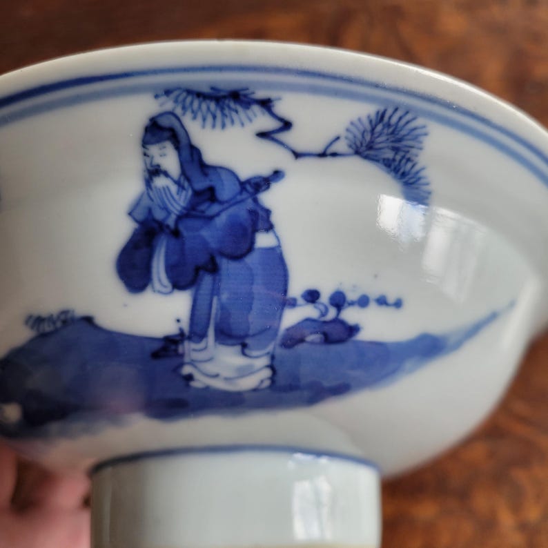 May include: A white porcelain bowl with a blue painted design. The bowl features a detailed illustration of a figure in traditional clothing, set against a landscape with trees. The rim is accented with a blue border. The bowl is elevated on a short, white pedestal.
