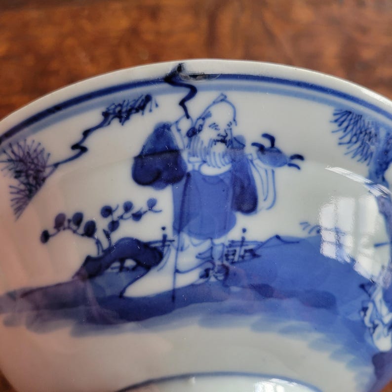 May include: A white and blue ceramic bowl with a detailed illustration of a man in a blue robe. The bowl's rim features a blue border with additional decorative elements. The artwork is in a traditional Asian style.
