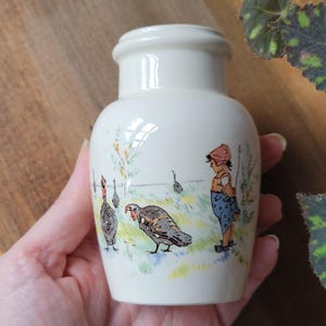 May include: A small, cream-colored ceramic jar with a smooth, rounded body and a short neck. The exterior features a colorful illustration of a person in a blue patterned outfit standing in a field with several turkeys among green and yellow grass.