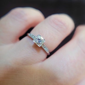 May include: A silver engagement ring with a square-shaped diamond set in a prong setting. The band is adorned with small diamonds.