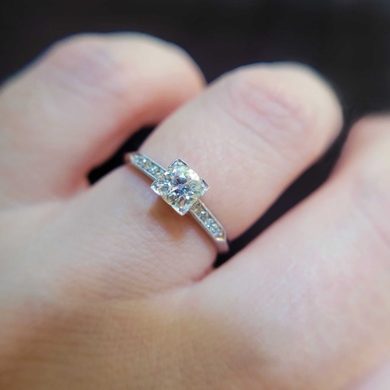 May include: A silver engagement ring with a square-shaped diamond set in a prong setting. The band is adorned with small diamonds.