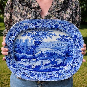 May include: An oval ceramic serving platter featuring a blue and white transferware pattern. The central scene depicts a rural landscape with a castle, a water mill, grazing cows, and figures on horseback. The border has a floral and leaf motif.