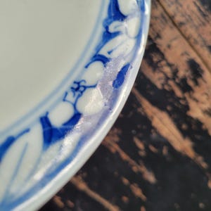 May include: Close-up of a white ceramic plate with a blue floral border design. The plate's edge is visible, showcasing the intricate blue and white pattern. The plate is partially visible, with a dark wooden surface in the background.