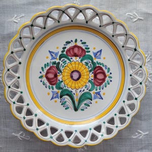 May include: A round ceramic plate with a scalloped, lattice-style openwork rim featuring yellow and green accents. The center displays a hand-painted floral motif with a large yellow sunflower, red blossoms, and green leaves on a white base.