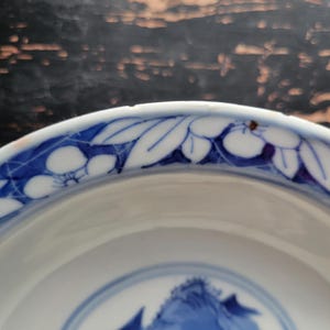 May include: A white ceramic bowl with a blue floral design around the rim and a blue mountain scene in the center. The bowl has a glossy finish and a slightly curved shape. The blue and white color scheme gives the bowl a classic, elegant look.