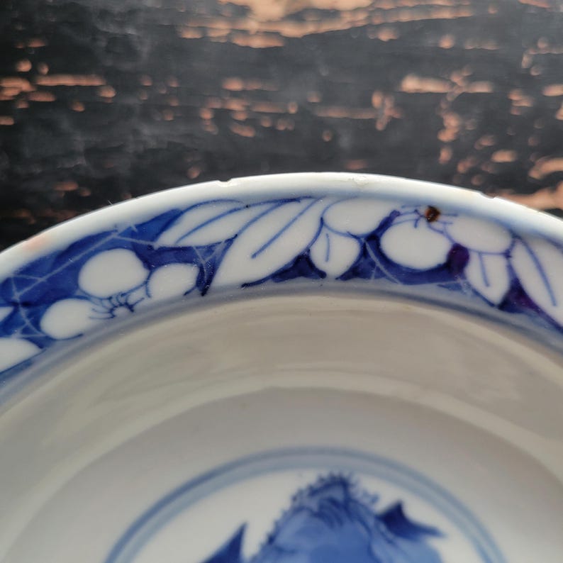 May include: A white ceramic bowl with a blue floral design around the rim and a blue mountain scene in the center. The bowl has a glossy finish and a slightly curved shape. The blue and white color scheme gives the bowl a classic, elegant look.