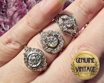 Pode incluir: Três anéis de diamante vintage com intrincados desenhos de filigrana. Os anéis são engastados em prata e apresentam diamantes redondos de corte brilhante. Os anéis são exibidos em uma mão que usa um tecido estampado de rosa e branco.