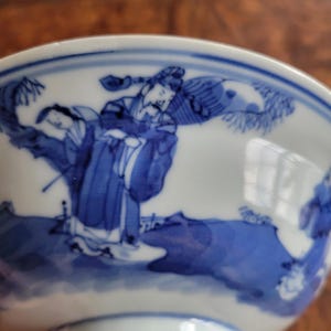 May include: A white ceramic bowl with a blue and white illustration of figures in traditional clothing. The bowl's design features a detailed landscape scene with figures, trees, and water. The bowl has a glossy finish.